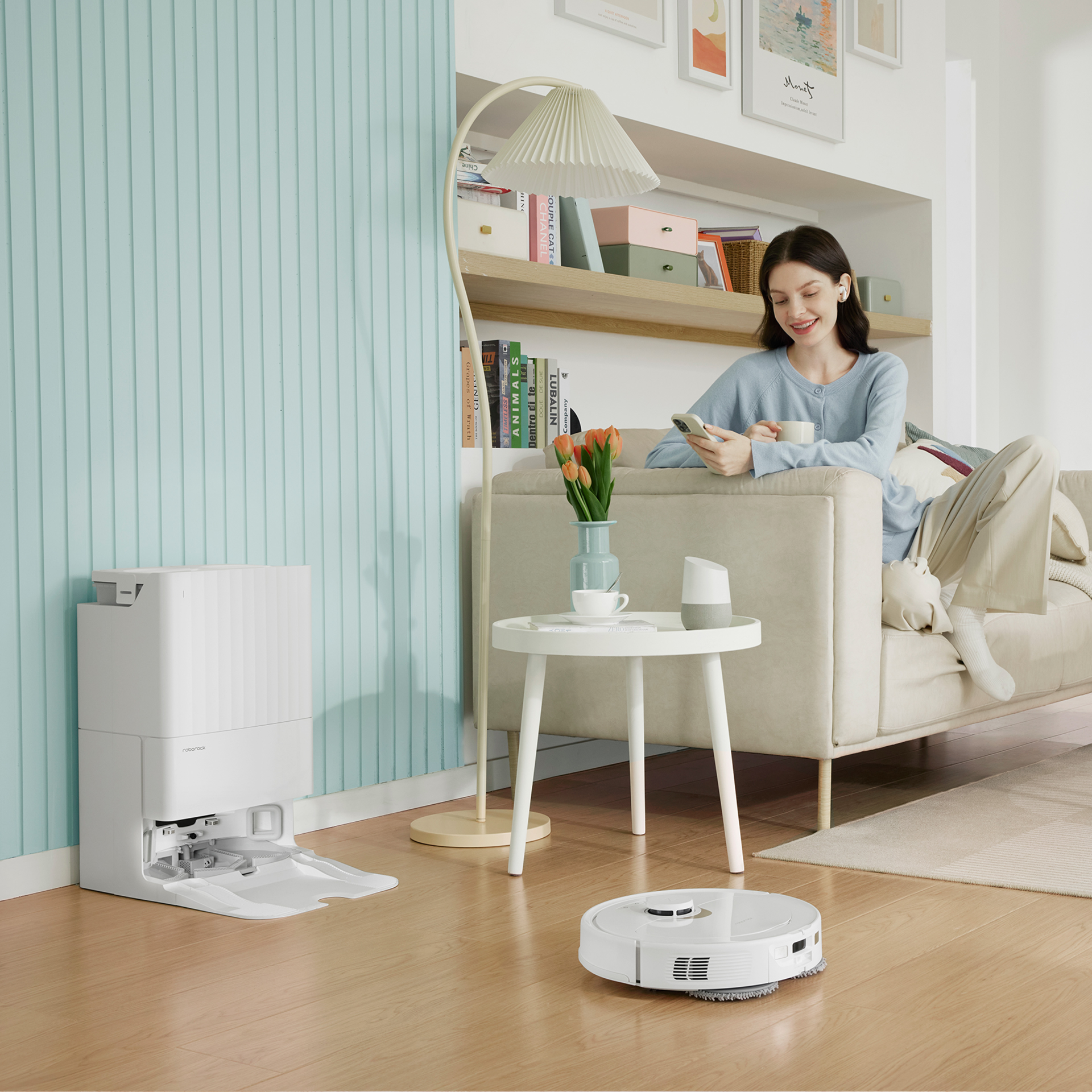 White Roborock robot vacuum docked in a white base station on a wooden floor. In the foreground, a hand holds a smartphone displaying a cleaning map on the Roborock app